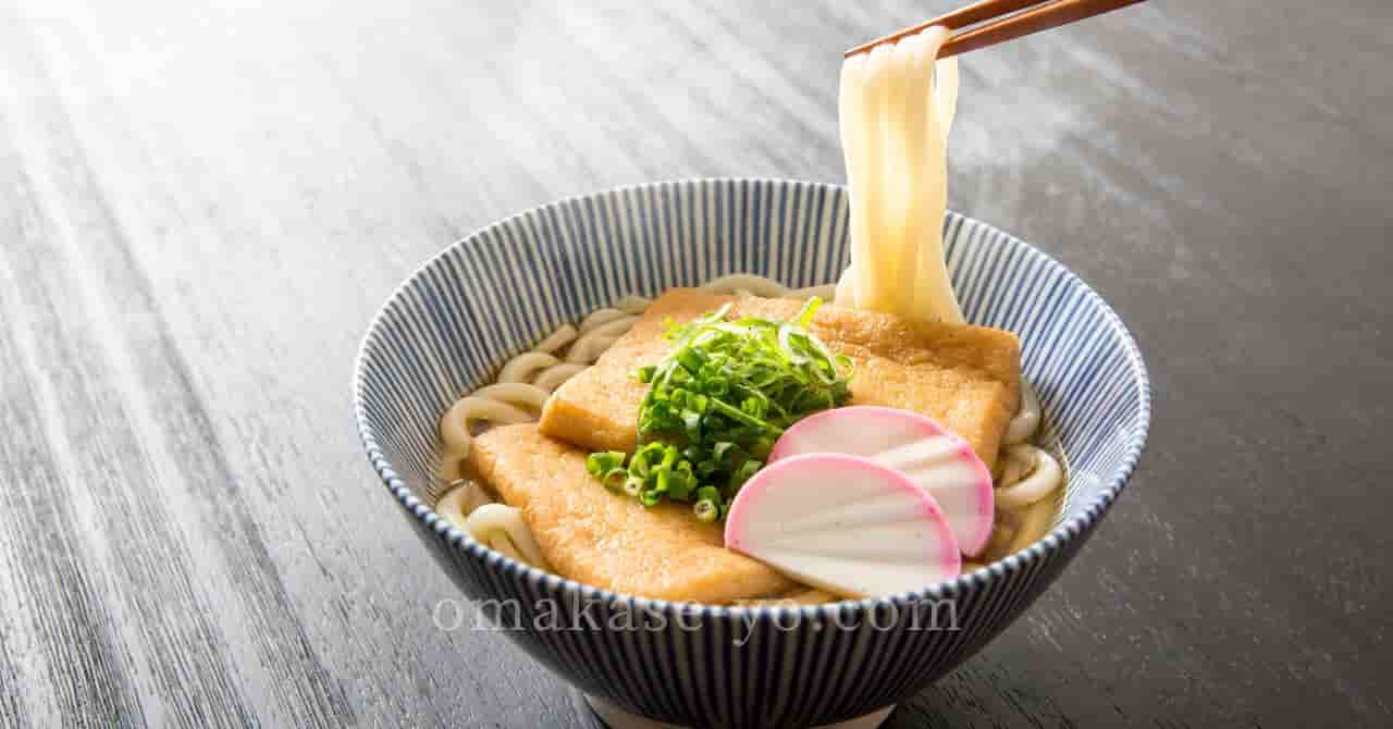 きつねうどんのどんぶり。上には刻みネギ、かまぼこ、油揚げが乗っており、箸でうどんを持ち上げているシーン。うなぎ以外の“う”のつく食材を紹介する記事に最適。